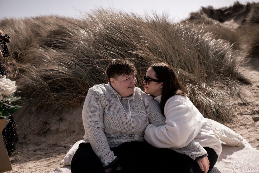 Two people sharing an intimate moment together while sitting against sand dunes on a beach.