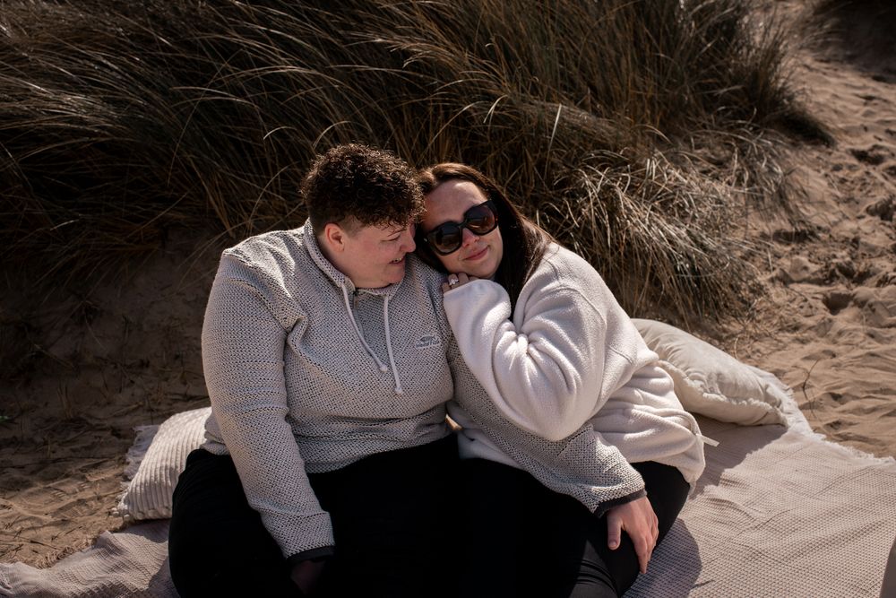 Couple sitting together on sandy beach dunes, wearing hoodies, enjoying a sunny day outdoors.