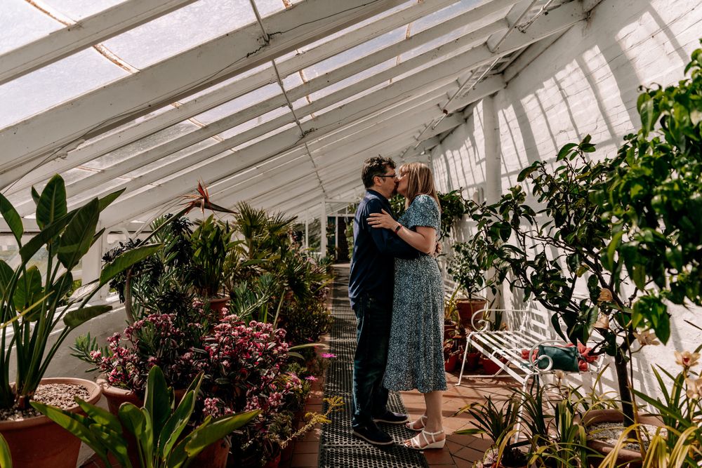 Couple embraces in a bright greenhouse filled with tropical plants and pink flowers under angled glass roof.