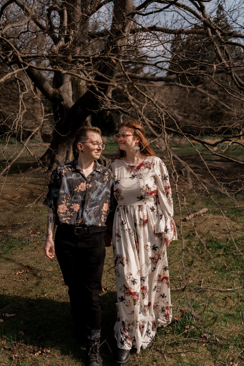 A couple in bloody zombie costumes pose together outdoors near bare trees in a Halloween themed photoshoot.