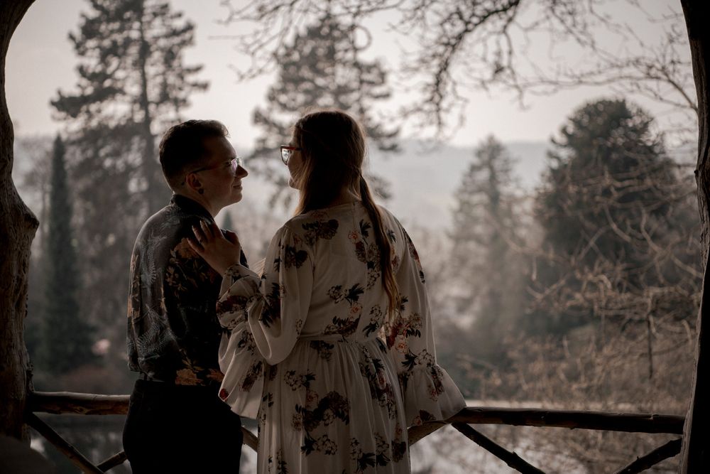 A romantic couple embraces in a rustic outdoor setting with evergreen trees and natural lighting in the background.