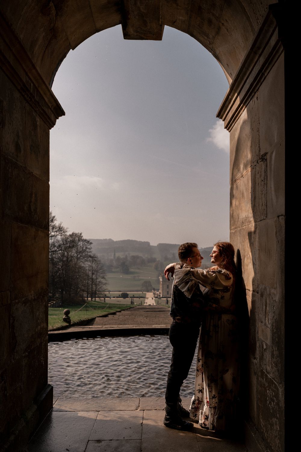 Two people silhouetted in a stone archway overlooking a misty countryside landscape at dusk.