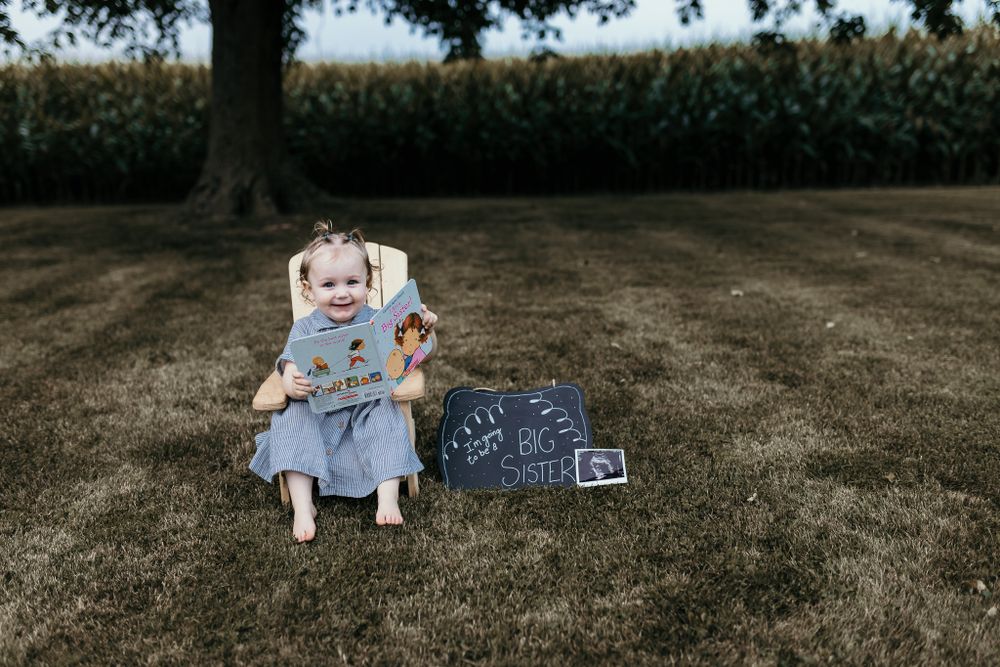 Child in blue dress sitting with pregnancy announcement chalkboard sign on grass.
