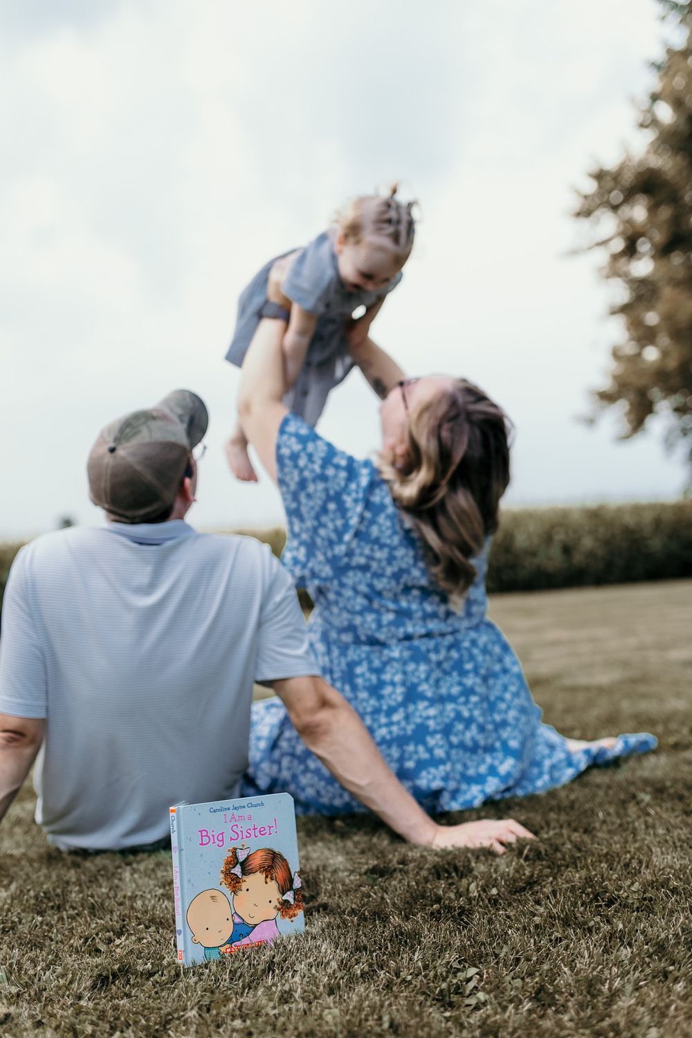 Family sitting on grass lifting toddler with pregnancy book in foreground.