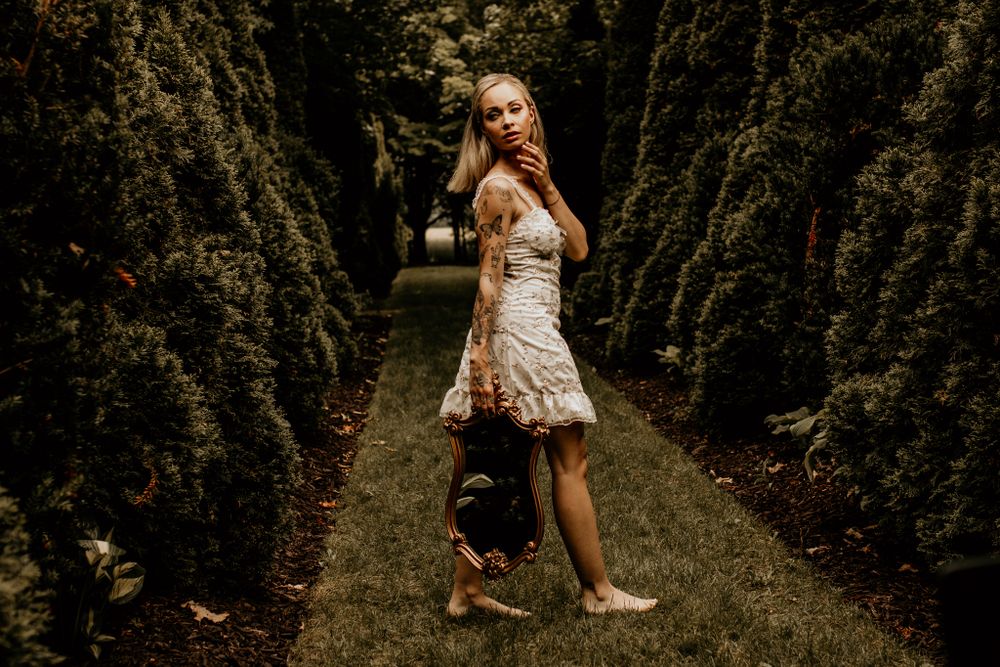 A woman in a flowing dress walks through a garden path lined with tall evergreen trees.