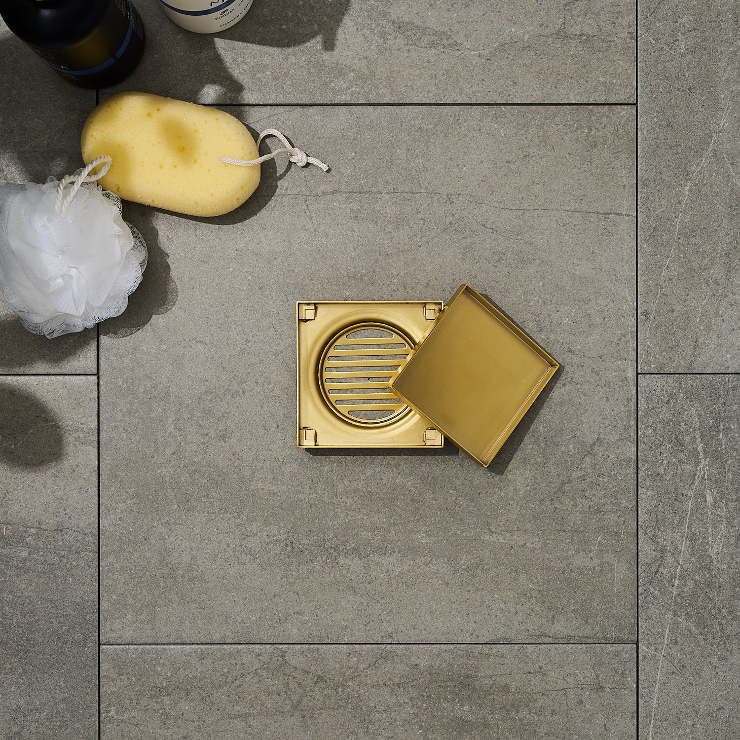Gold square grates sitting on tiles with bath sponge, luffa, and shampoo