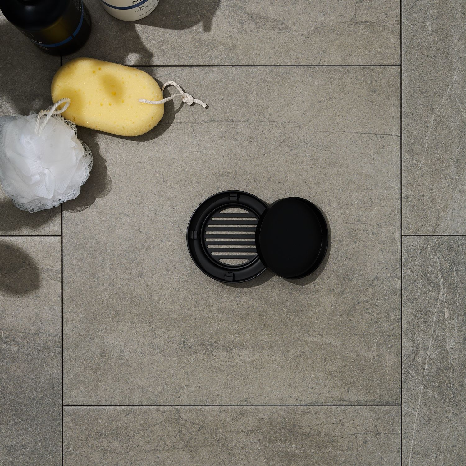 Black circular grates sitting on tiles with bath sponge, luffa, and shampoo