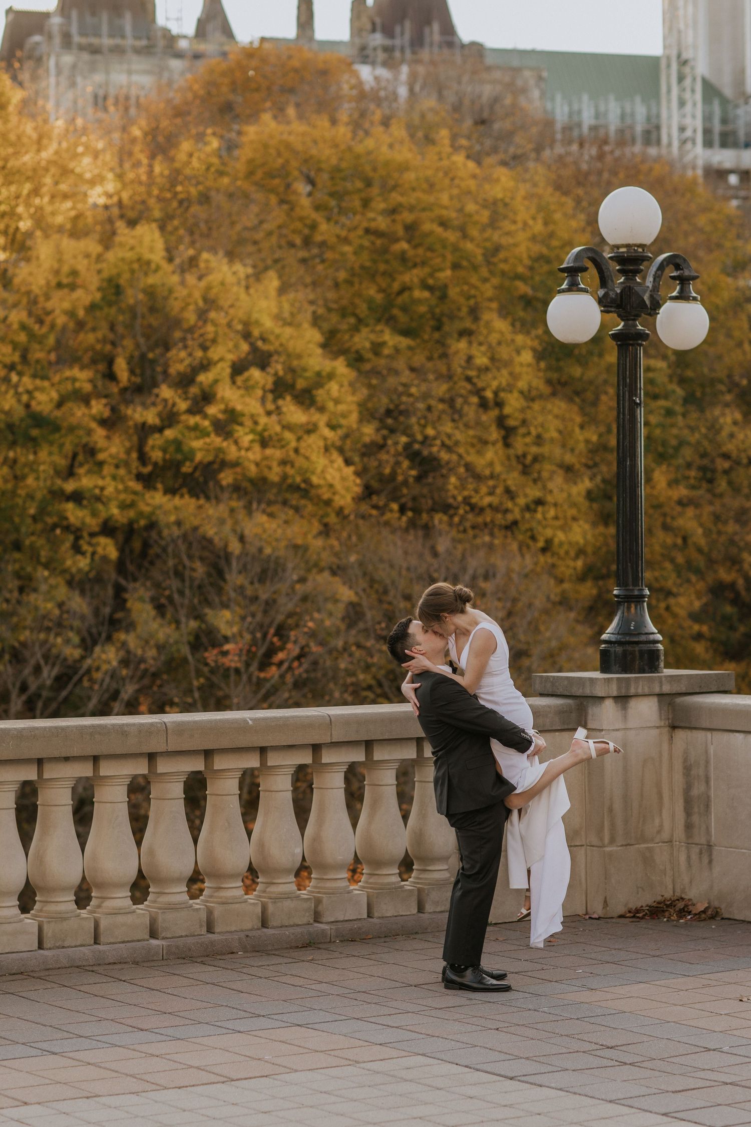 Wedding couple shares tender moment on stone balcony with autumn foliage backdrop.