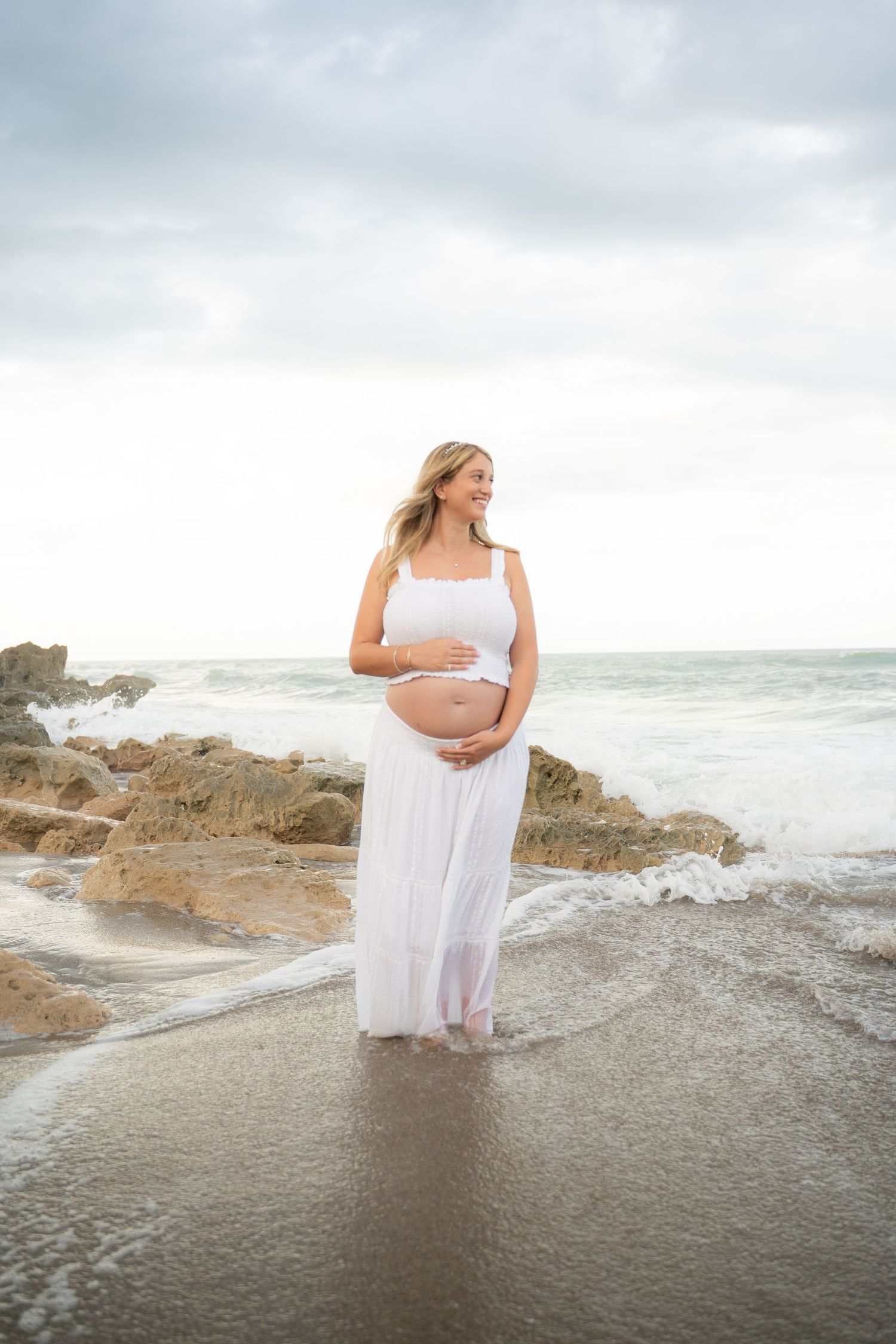 Pregnant woman in white crop top and flowing skirt standing barefoot on rocky beach with ocean waves.