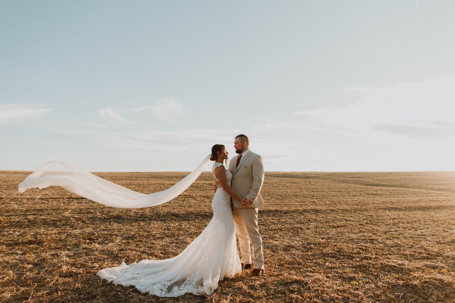 A couple in wedding attire stands in a golden field as the bride's veil flows dramatically in the wind.