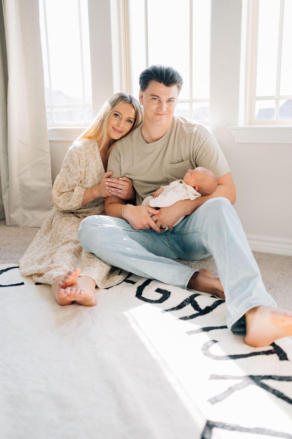 A couple sits in a bright room with their newborn baby on a carpeted floor beside decorative text.