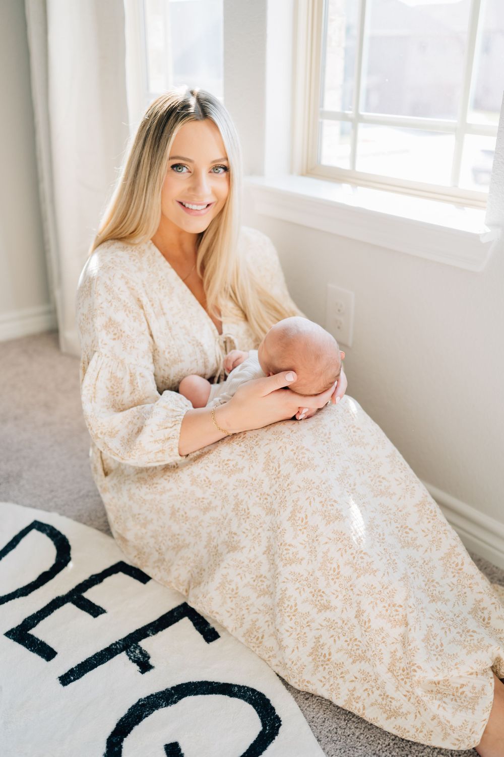 A woman in a lace dress sits by a window holding a newborn baby on a carpeted floor.