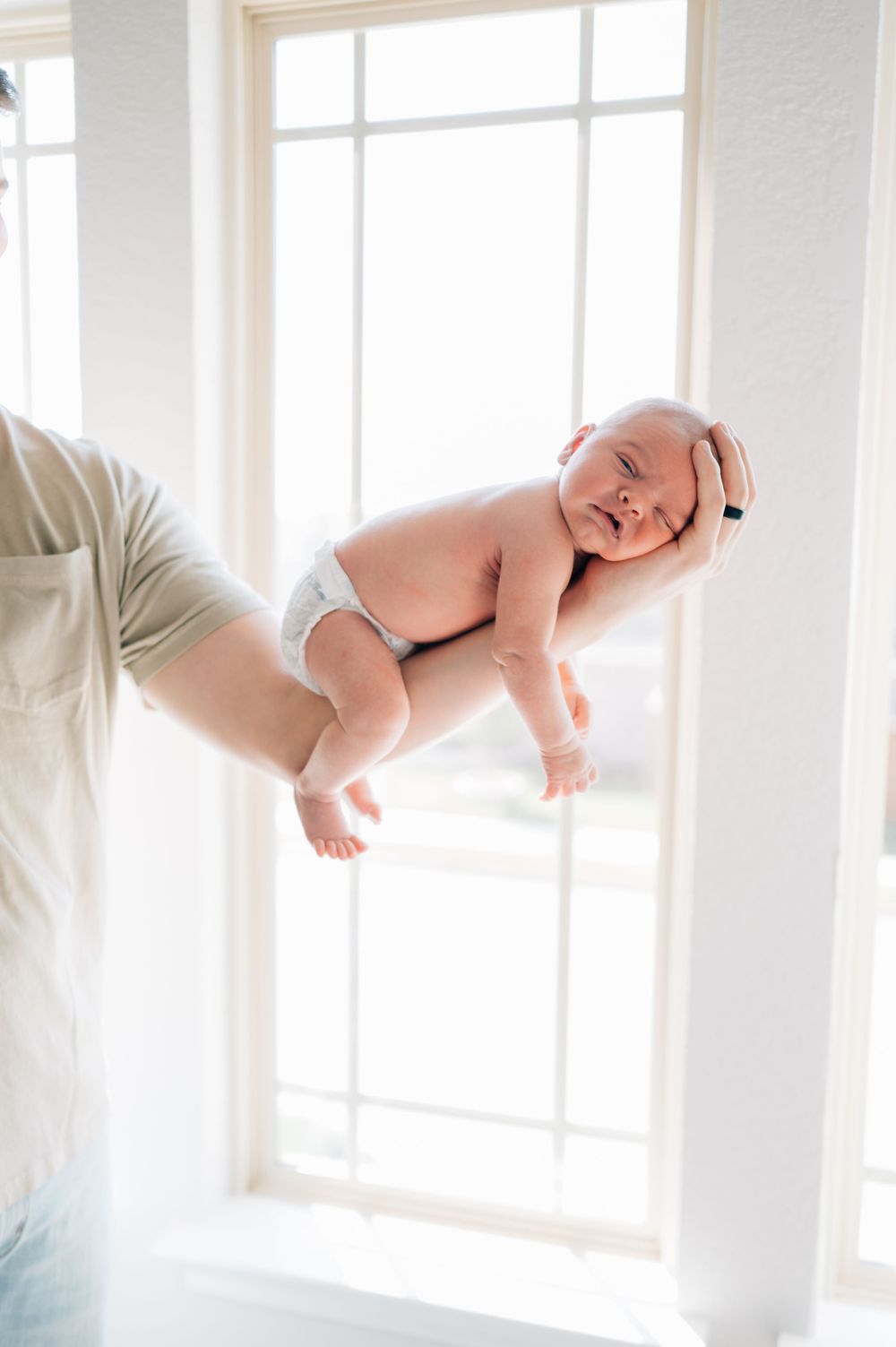 Smiling baby in diaper held up against bright window with sunlight streaming in.