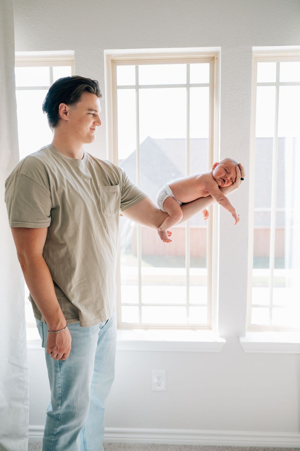 Person in grey t-shirt and jeans holding infant against sunlit window background.
