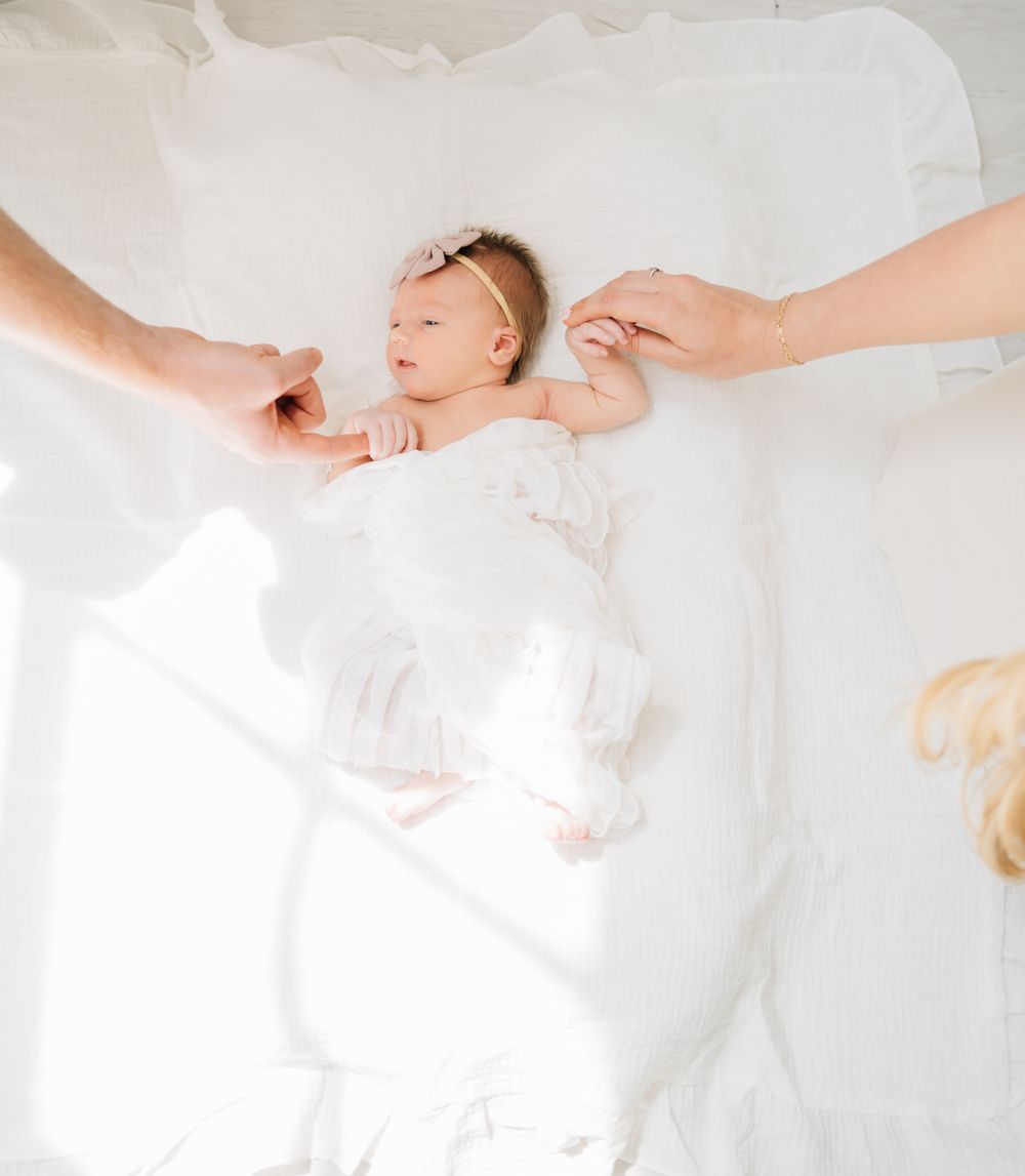 Newborn baby sleeping peacefully on white bedding with parents' hands reaching out.