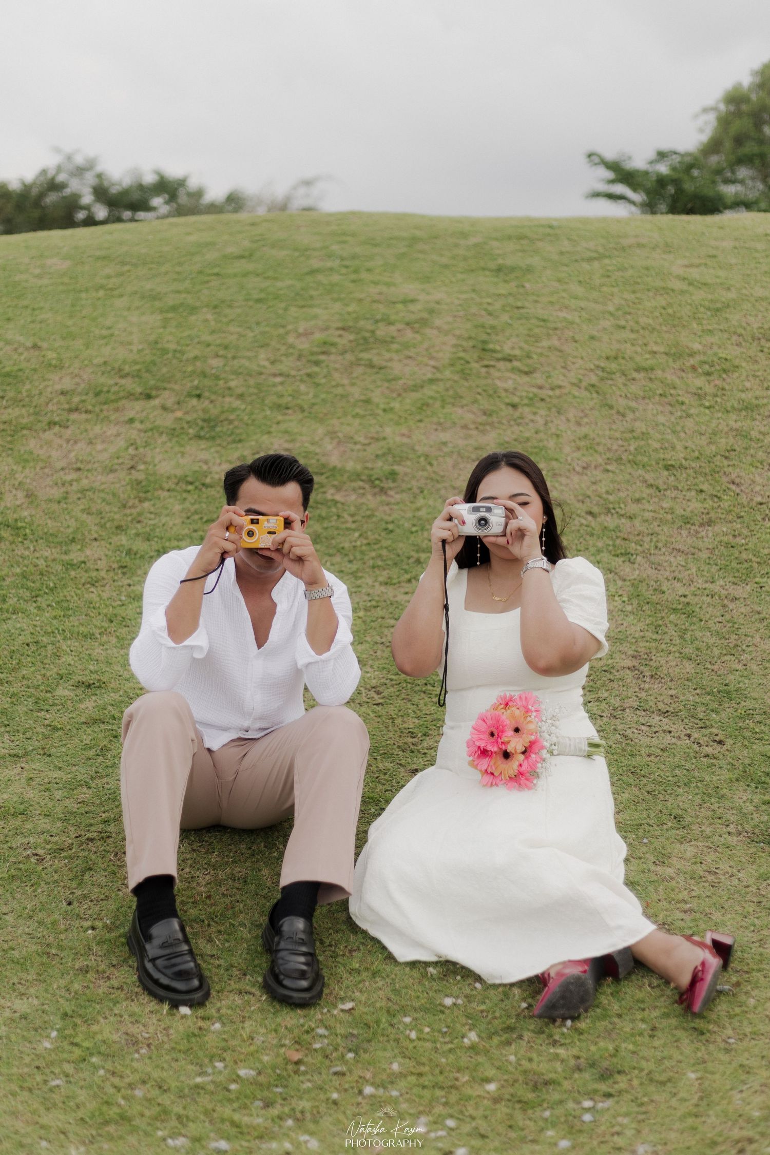 A couple in white attire share tender moments while holding flowers in an outdoor photoshoot.