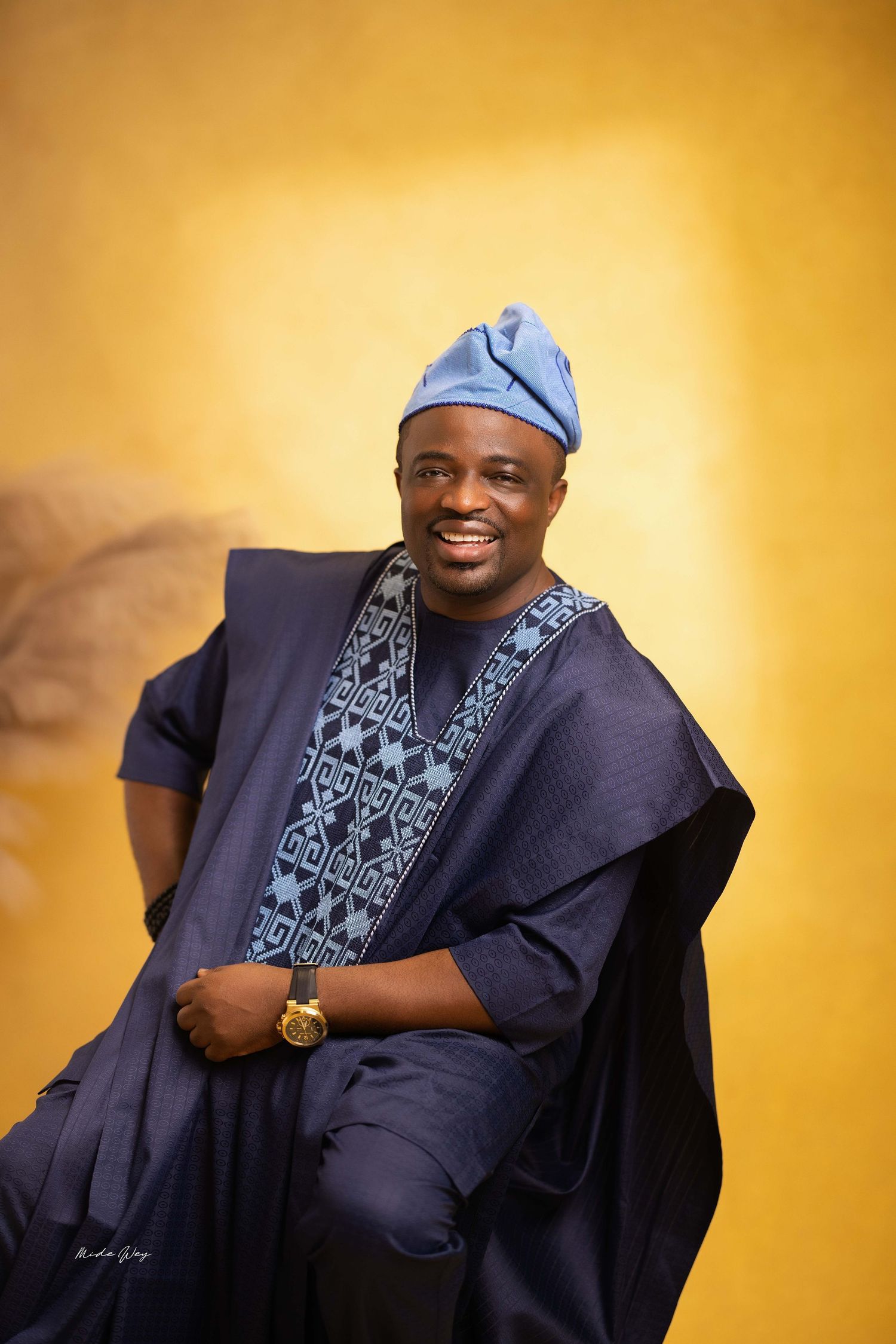 A person wearing traditional African attire and blue cap smiles warmly against a golden yellow background.
