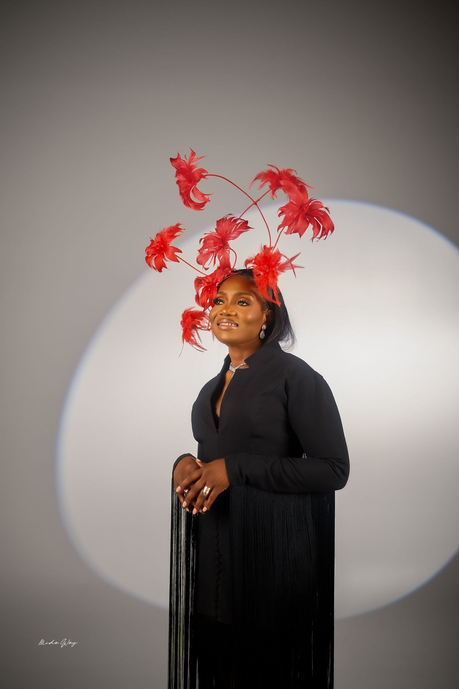 A performer in a black outfit with vibrant red feathers in their hair poses expressively against a light background.