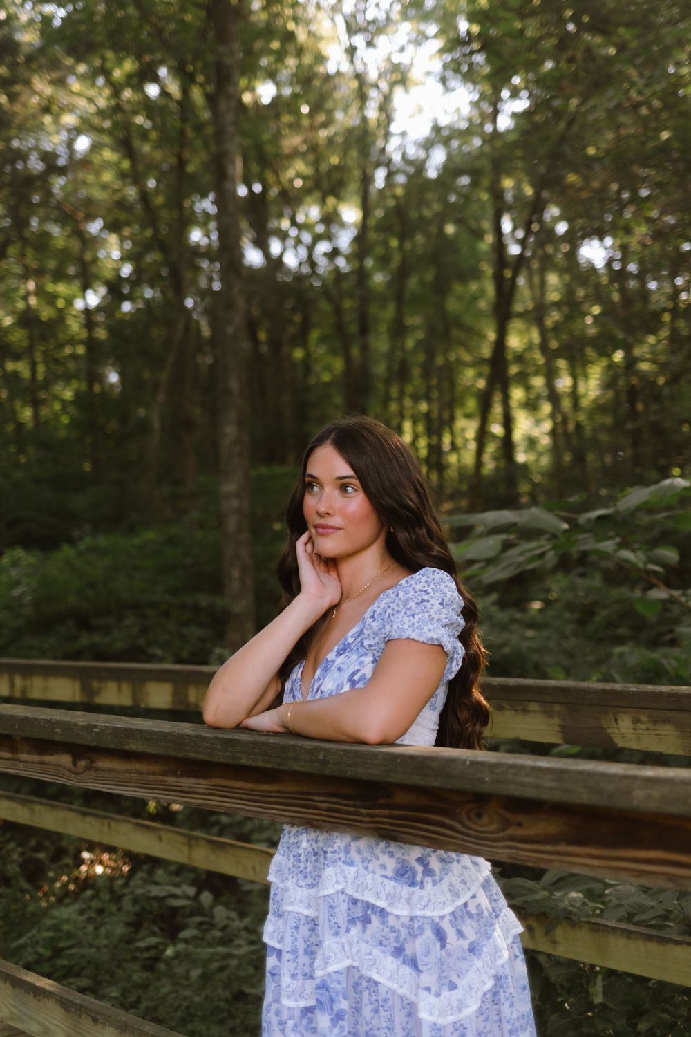 Person in a blue and white lace dress poses thoughtfully on a wooden railing in a wooded forest setting at sunset.
