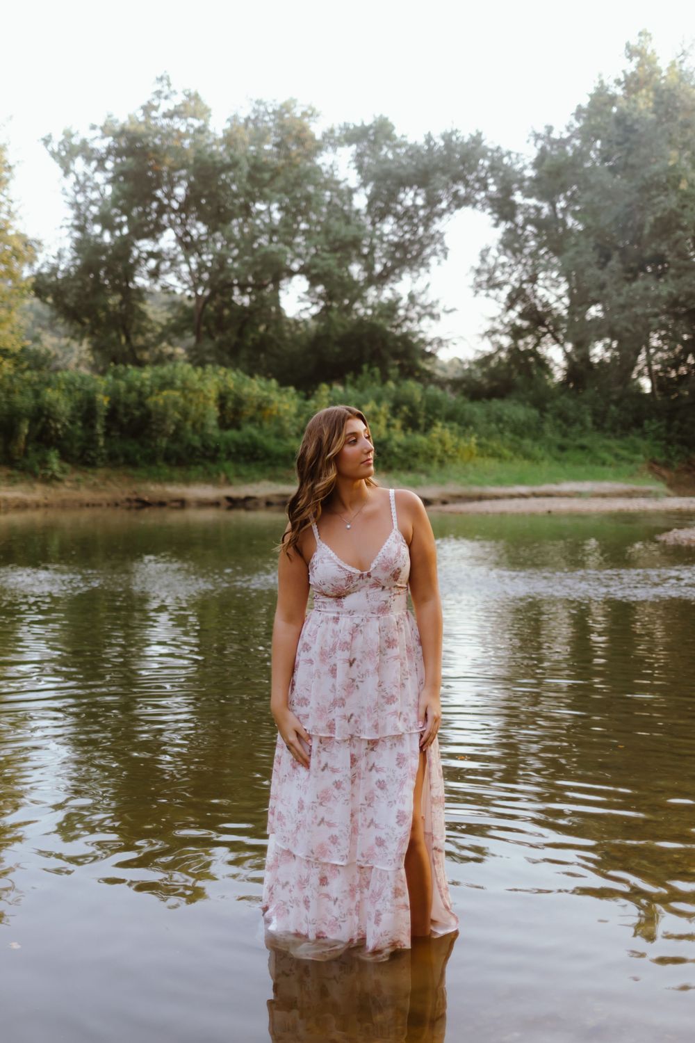 Person in flowing floral maxi dress stands in shallow water by wooded riverbank during golden hour.