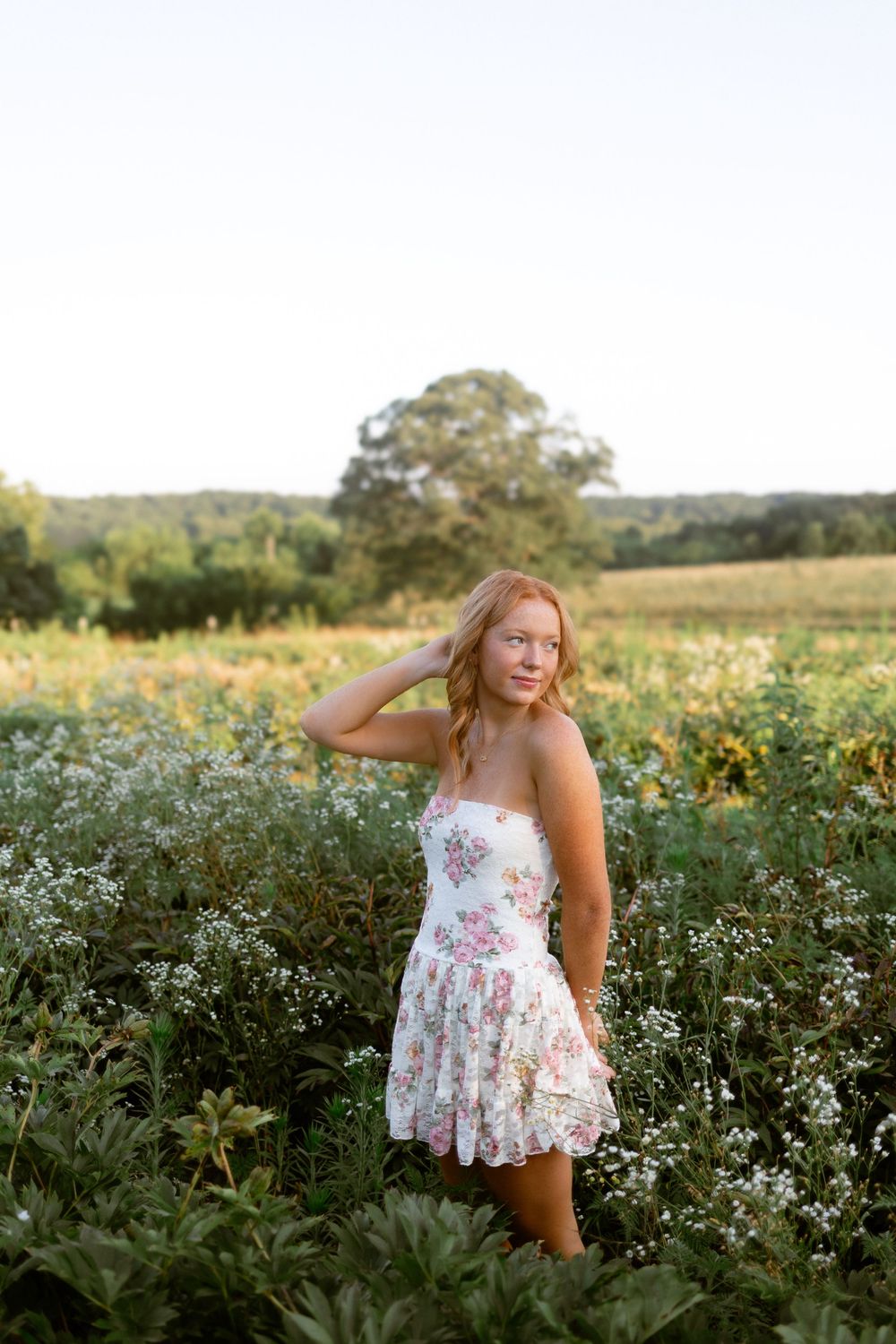 A person in a floral dress poses playfully in a field of white wildflowers and sunflowers on a sunny summer day.