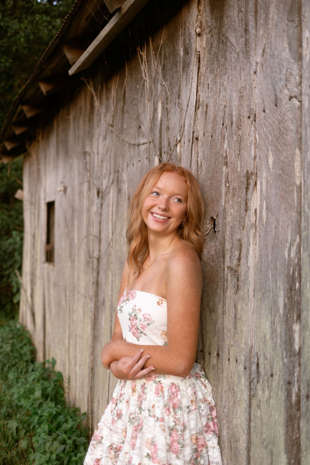 A figure in a floral white dress poses against a rustic wooden barn wall in a series of outdoor portraits.