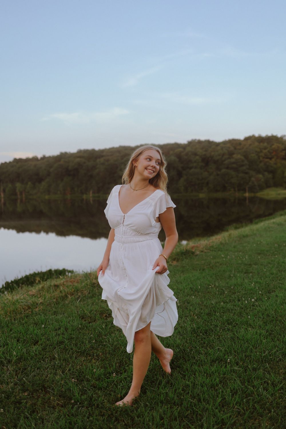 A woman in a white dress twirls playfully in a grassy field near a calm lake at sunset.