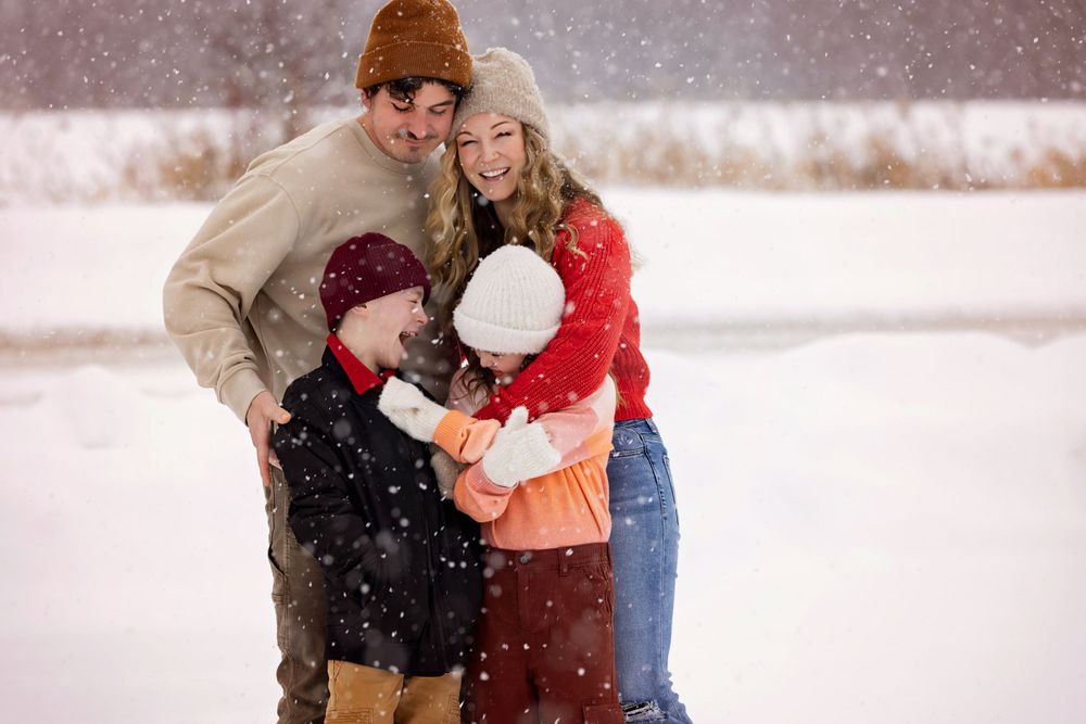 Family cuddles together in the snow wearing winter clothing during gentle snowfall.