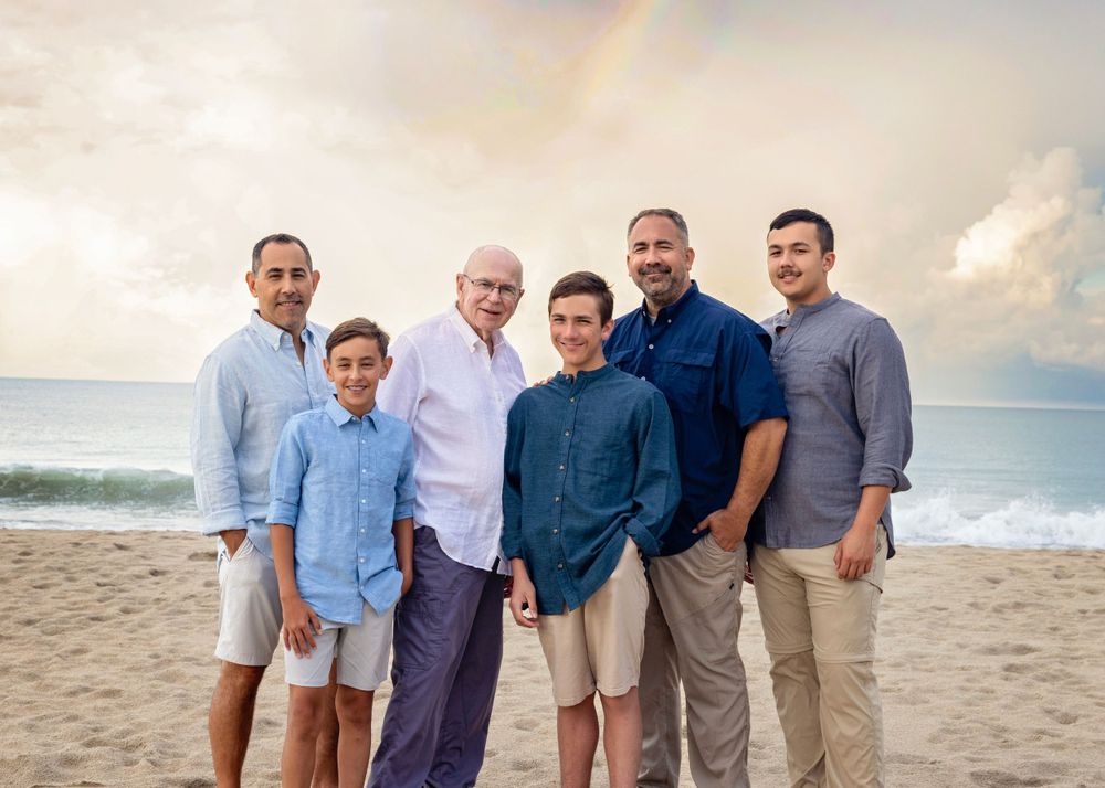 A multi-generational family group poses together on a beach at sunset with a rainbow in the background.
