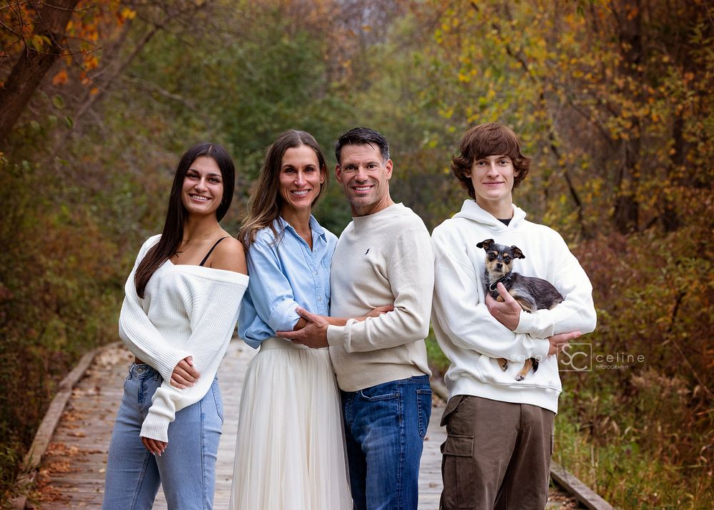 A family group poses together on a wooden bridge surrounded by autumn foliage while holding a small dog.