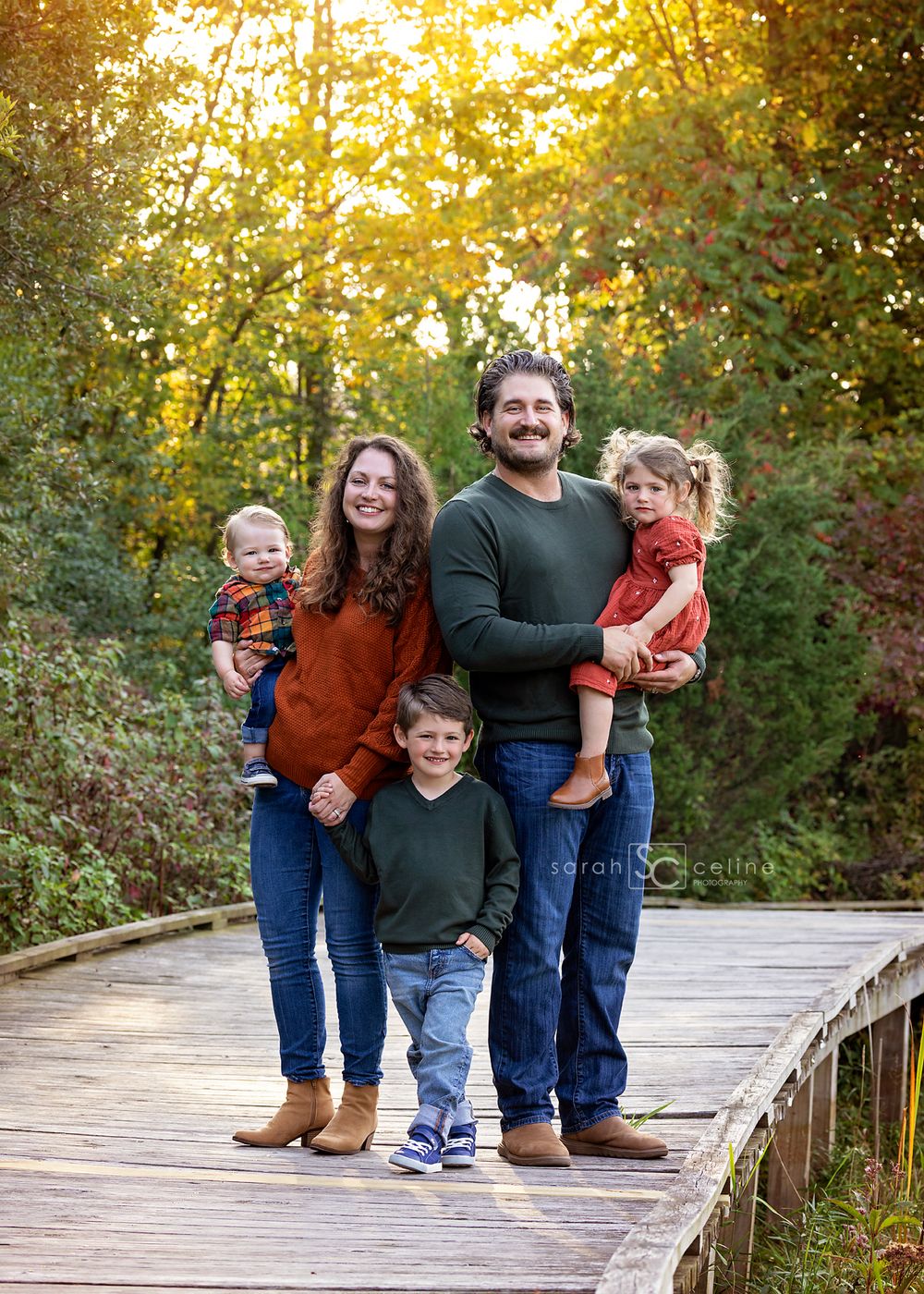 A family stands together on a wooden boardwalk surrounded by autumn foliage during a fall photo session.