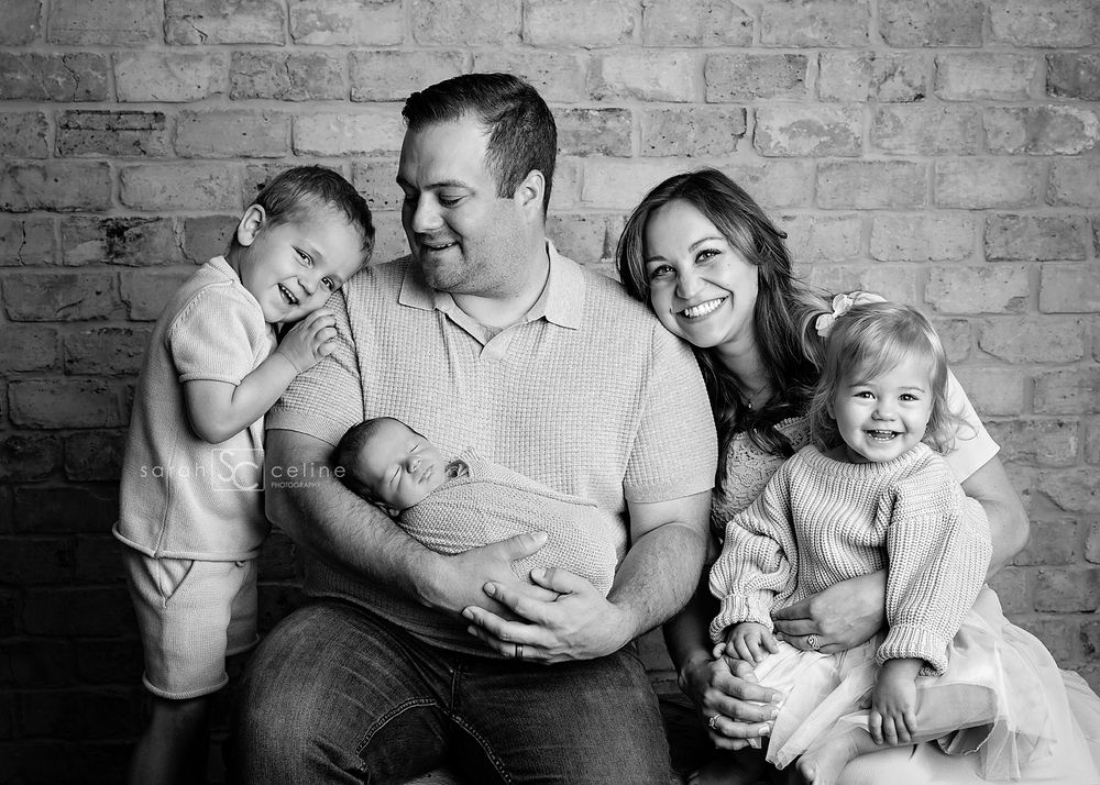 Black and white portrait of a casual family sitting together with three children against a brick wall.