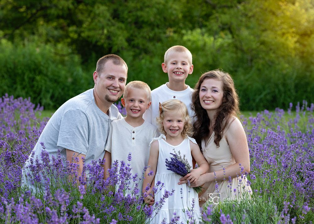 Family of five smiling in a vibrant lavender field, with lush green trees in the background on a sunny day.