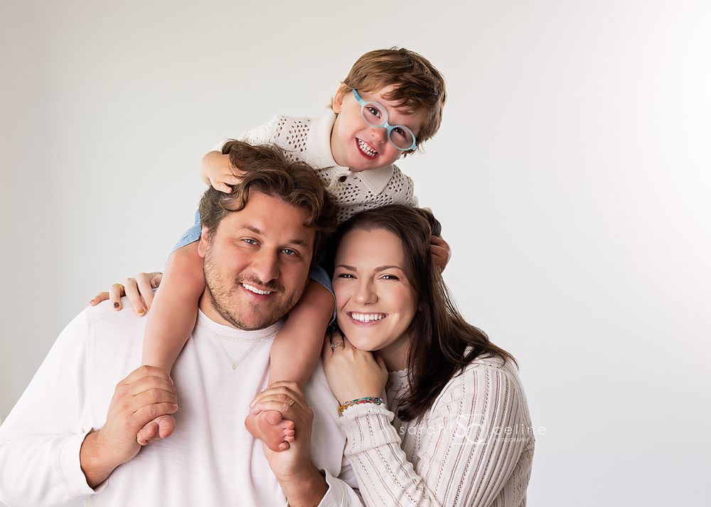 Happy family portrait of smiling parents with young child on dad's shoulders against white background.