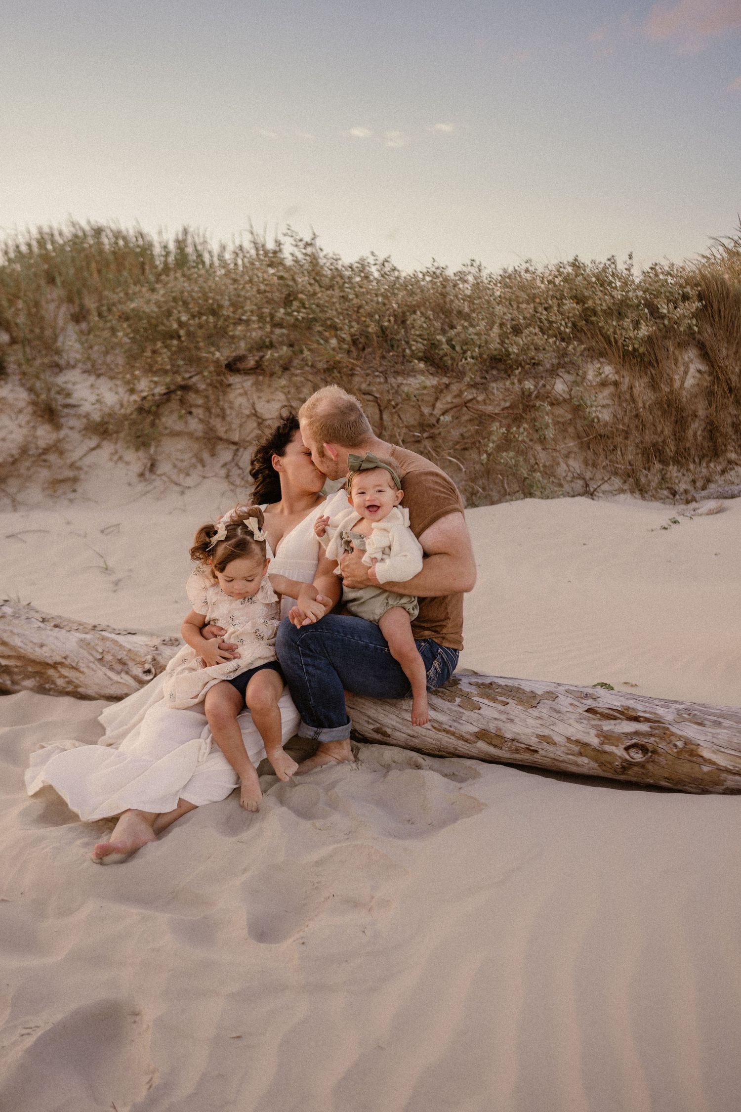 Family shares tender moment while sitting together on driftwood at sandy beach during sunset.