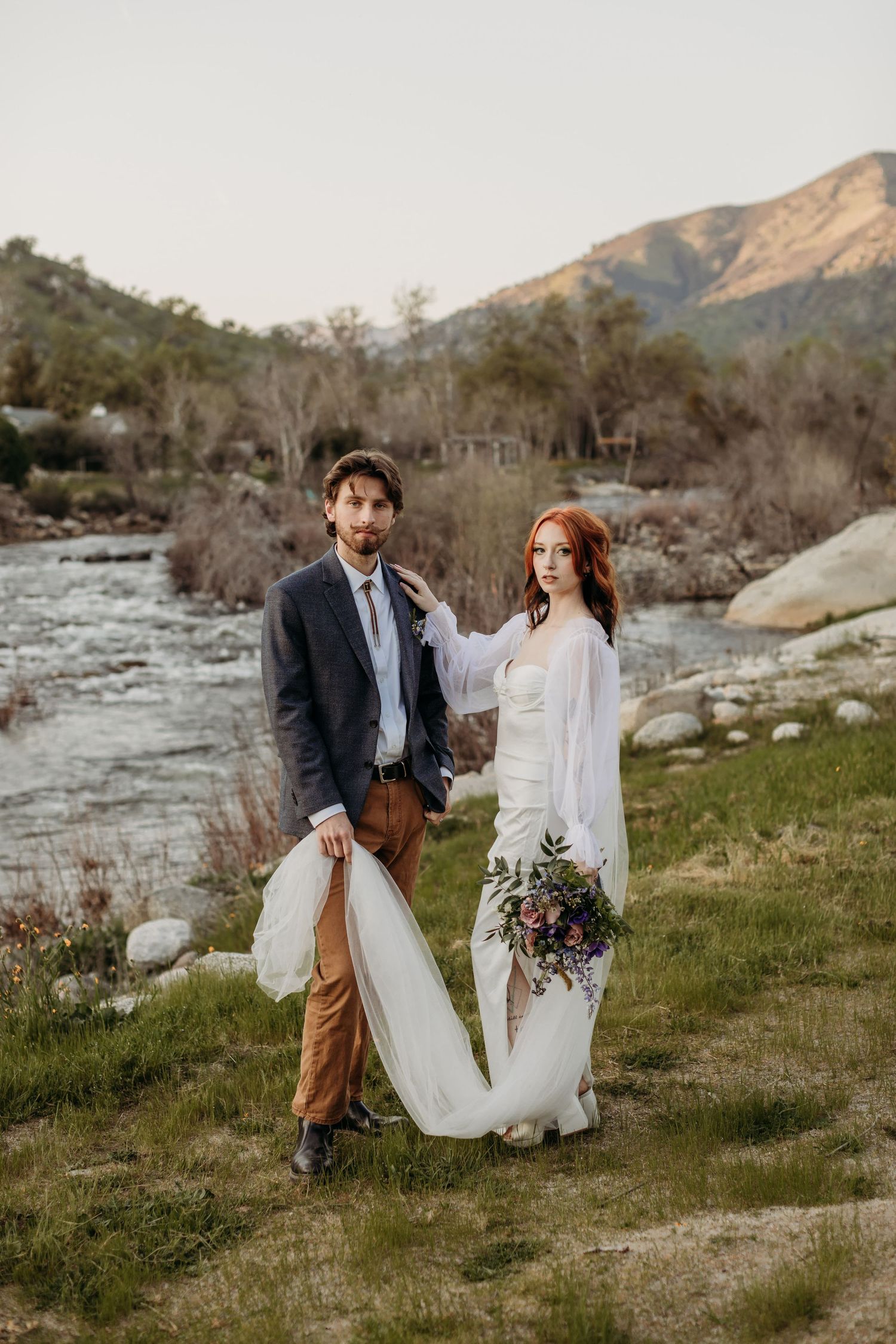 Couple in wedding attire poses together by mountain river with flowing white dress.