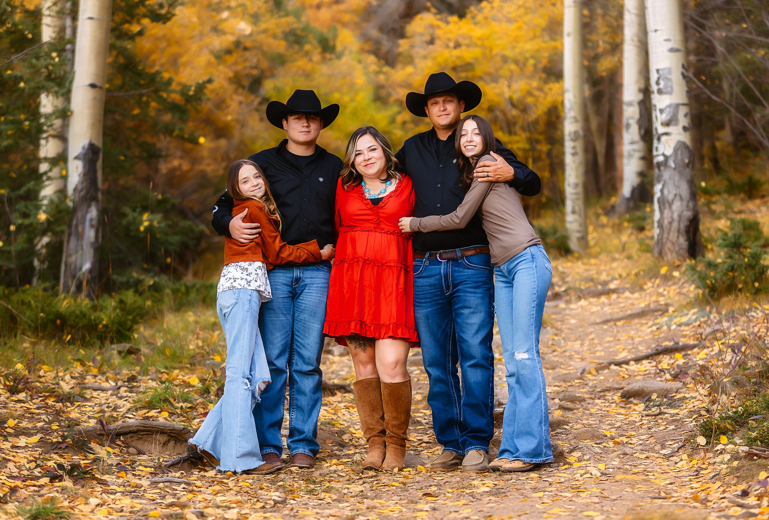 A group in western attire poses together on a woodland trail surrounded by golden autumn foliage and birch trees.