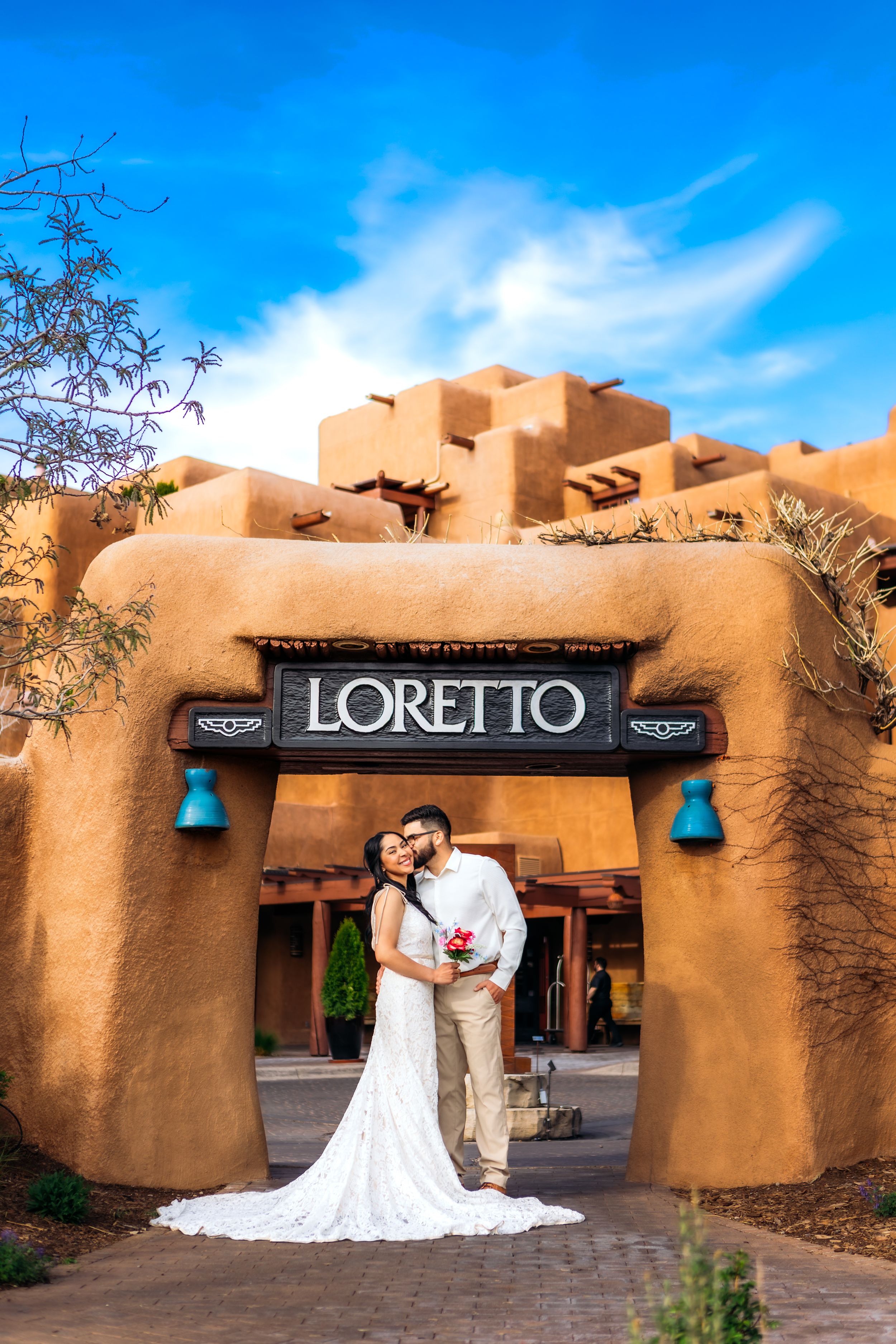 A couple in wedding attire embraces beneath the adobe archway entrance of the Loretto building against a bright blue sky.