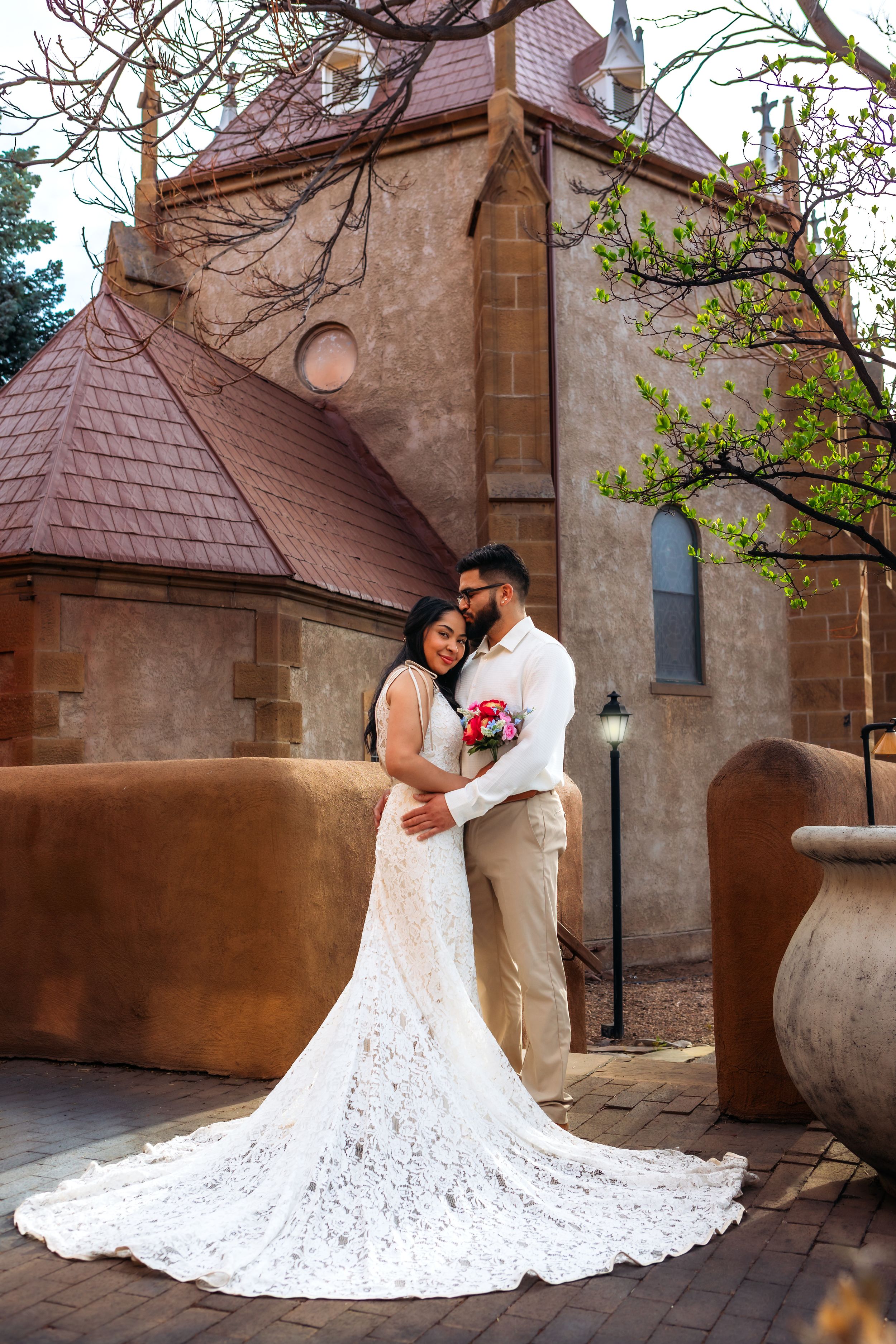 A wedding couple embraces in a romantic moment against a rustic stone building wall with a flowing white lace dress train.