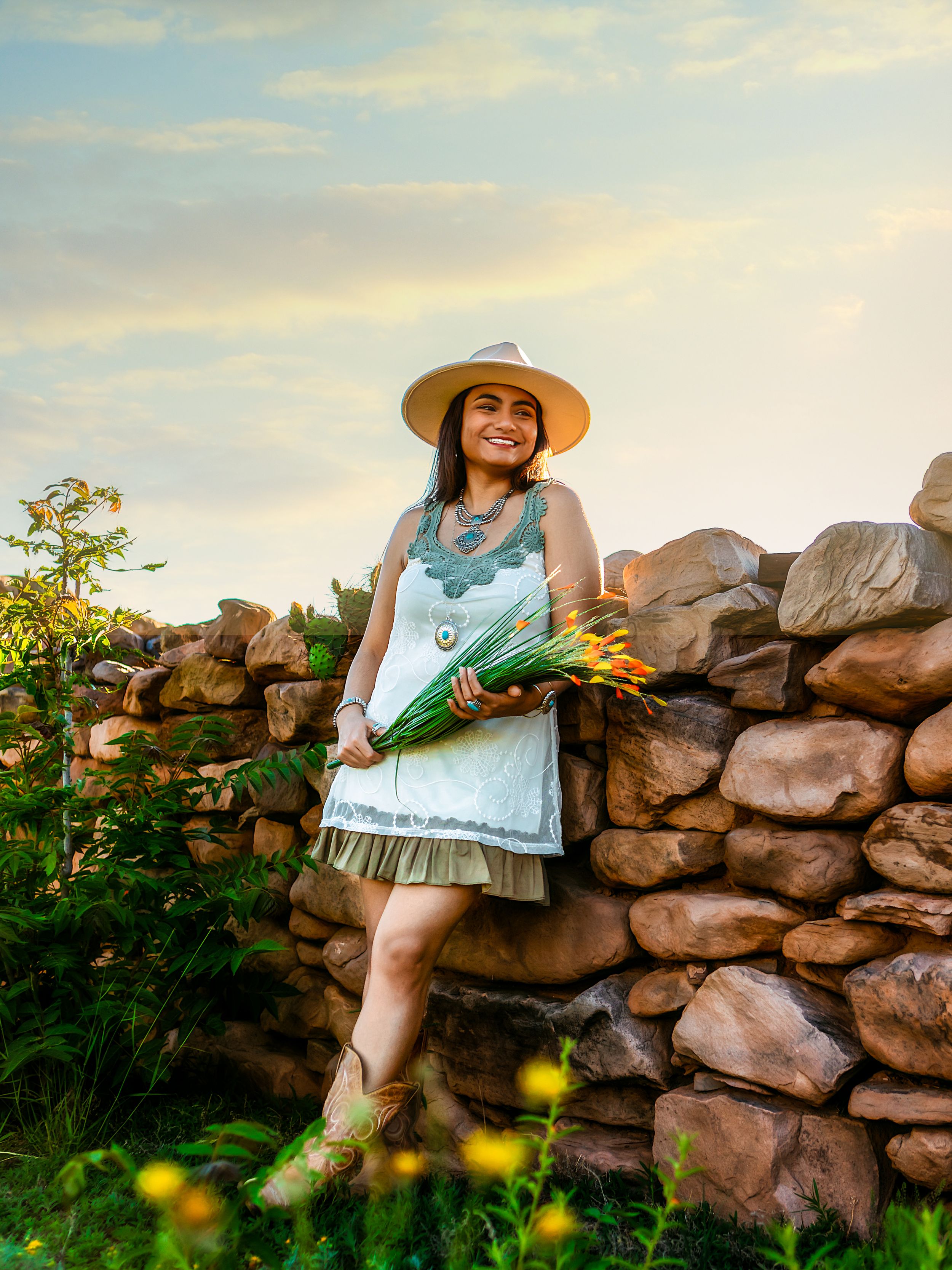 A person in a sun hat and white dress sits atop a rustic stone wall surrounded by greenery in a summer garden setting.