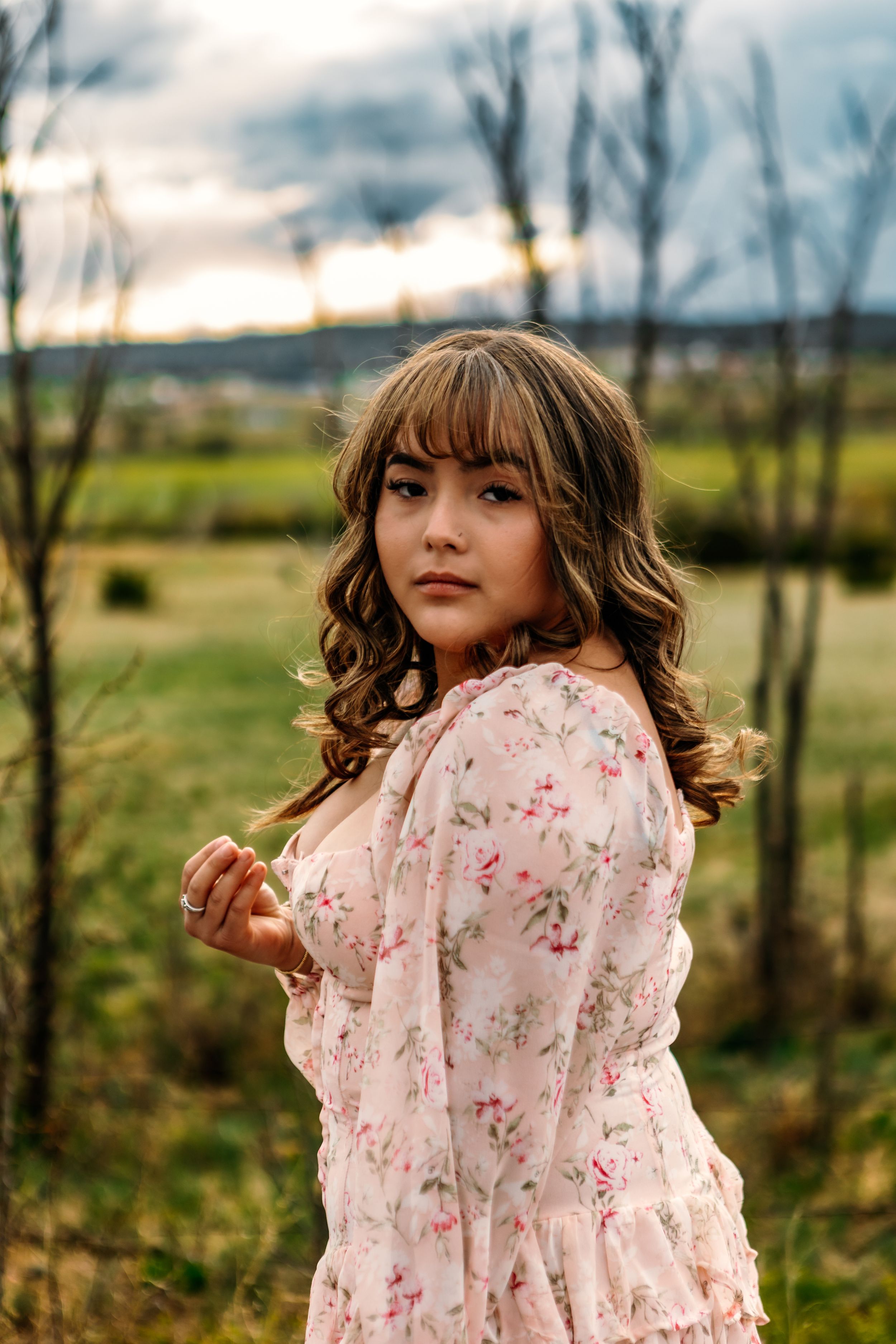 A person in a floral pink dress poses thoughtfully in a rural field at sunset with trees in the background.