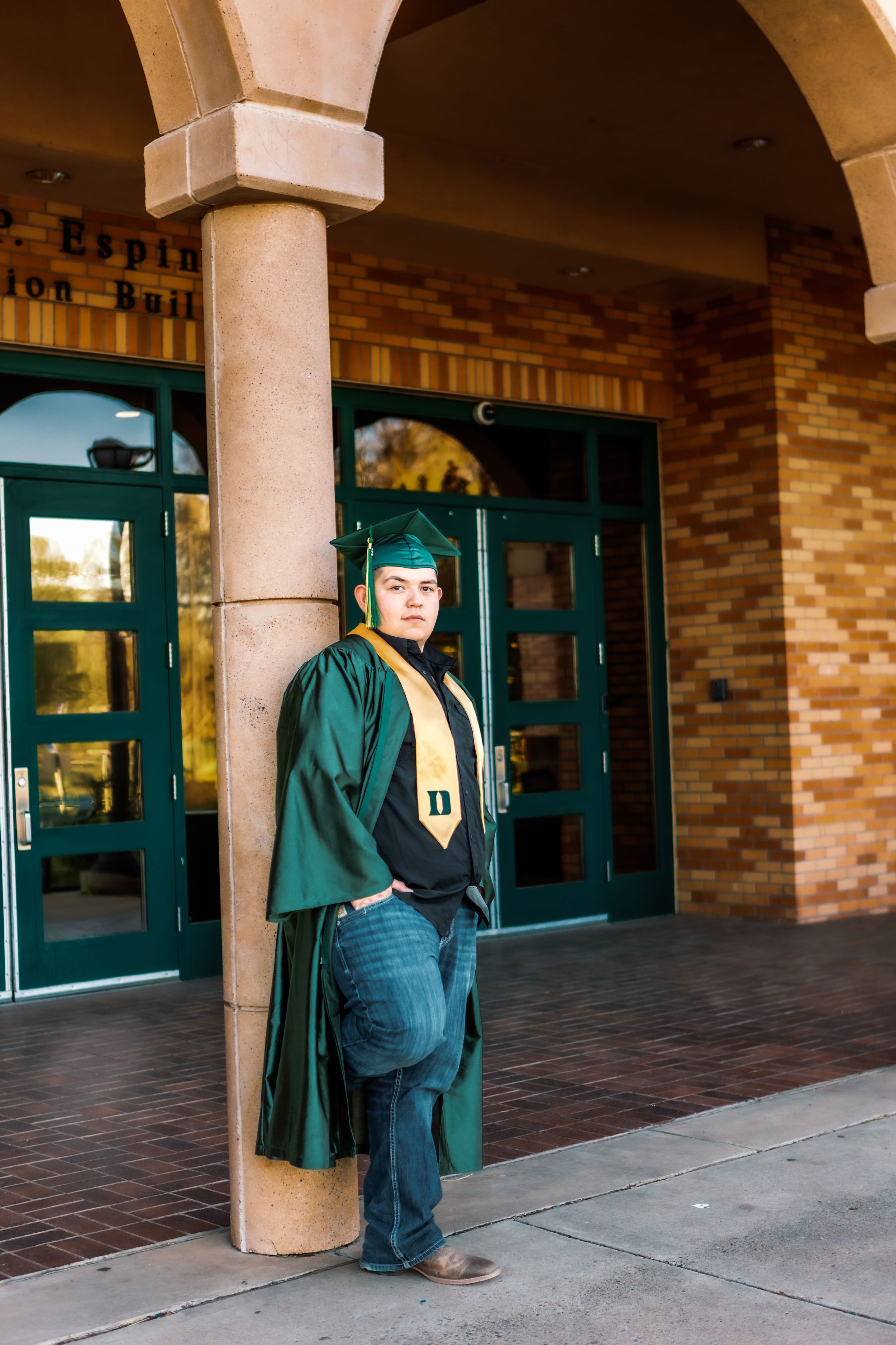 A graduate in dark green cap and gown stands in an archway entrance with brick walls and green doors.