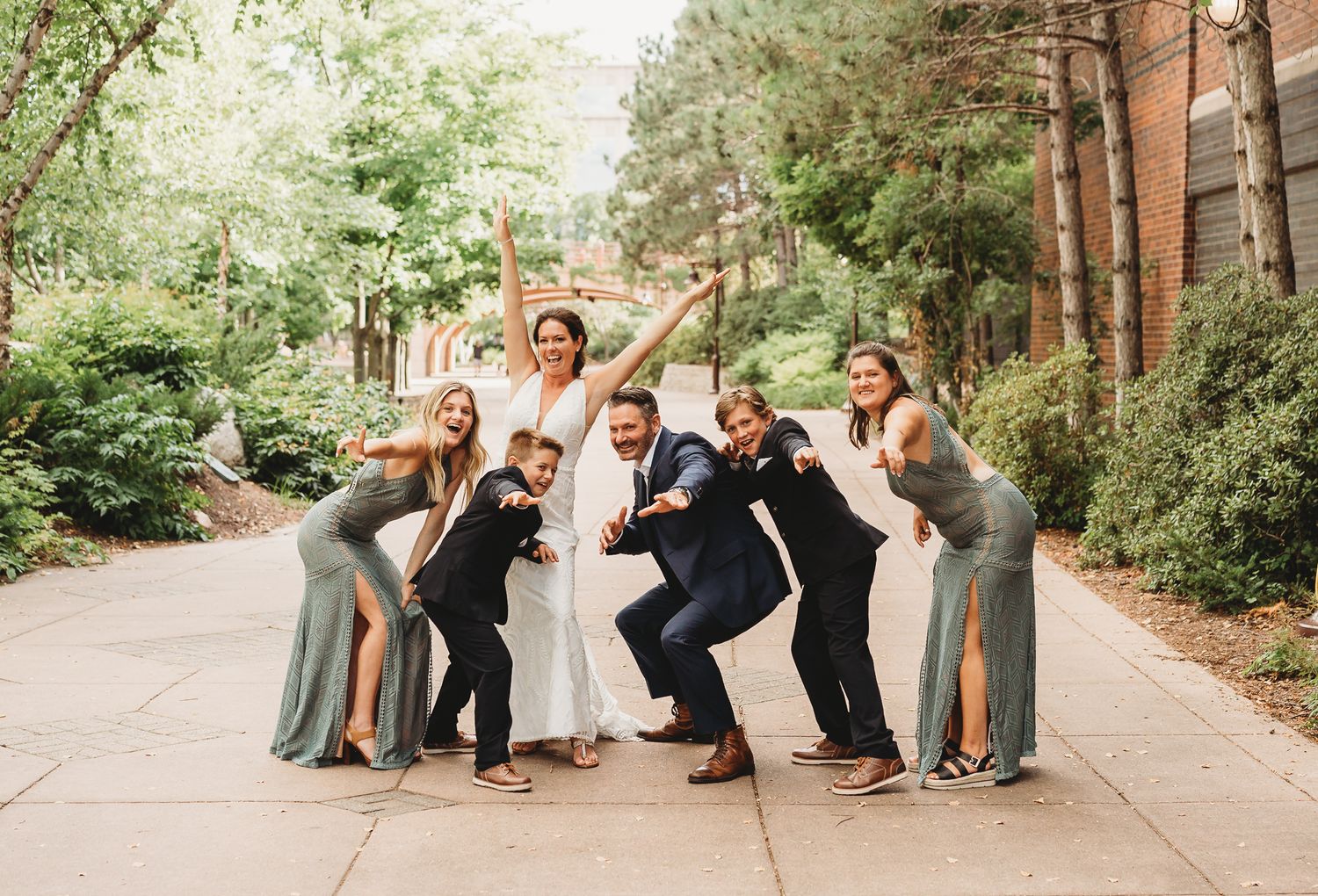 Wedding party poses playfully on a tree-lined path in coordinated formal attire with bridesmaids in gray dresses.