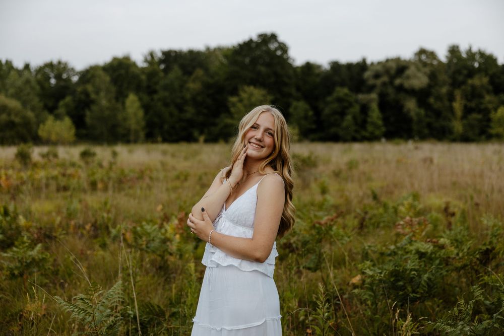 A young woman in a white dress stands in a field with trees in the background. Fort Worth Texas senior photographer.
