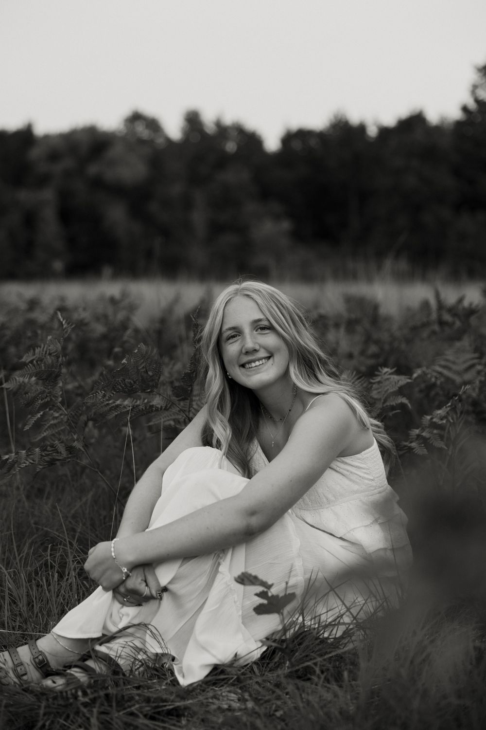 A young woman with long blonde hair sits in a grassy field, smiling and embracing herself. Fort Worth Texas senior photographer.