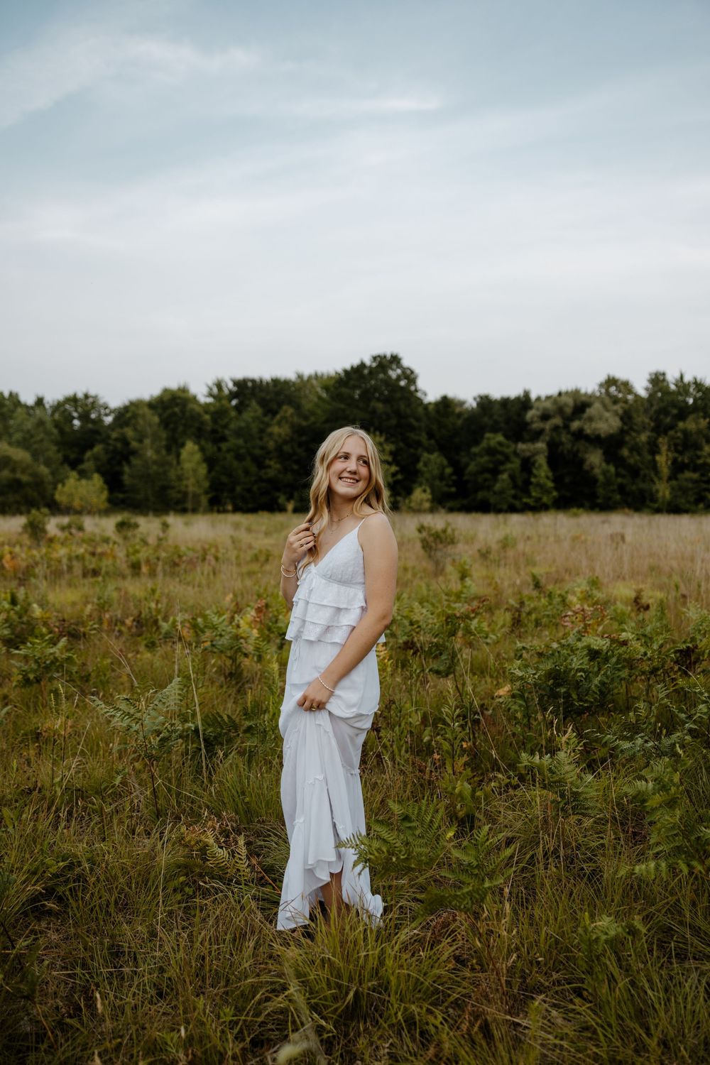 A young woman in a white dress stands in a grassy field surrounded by trees on a sunny day. Fort Worth Texas senior photographer.
