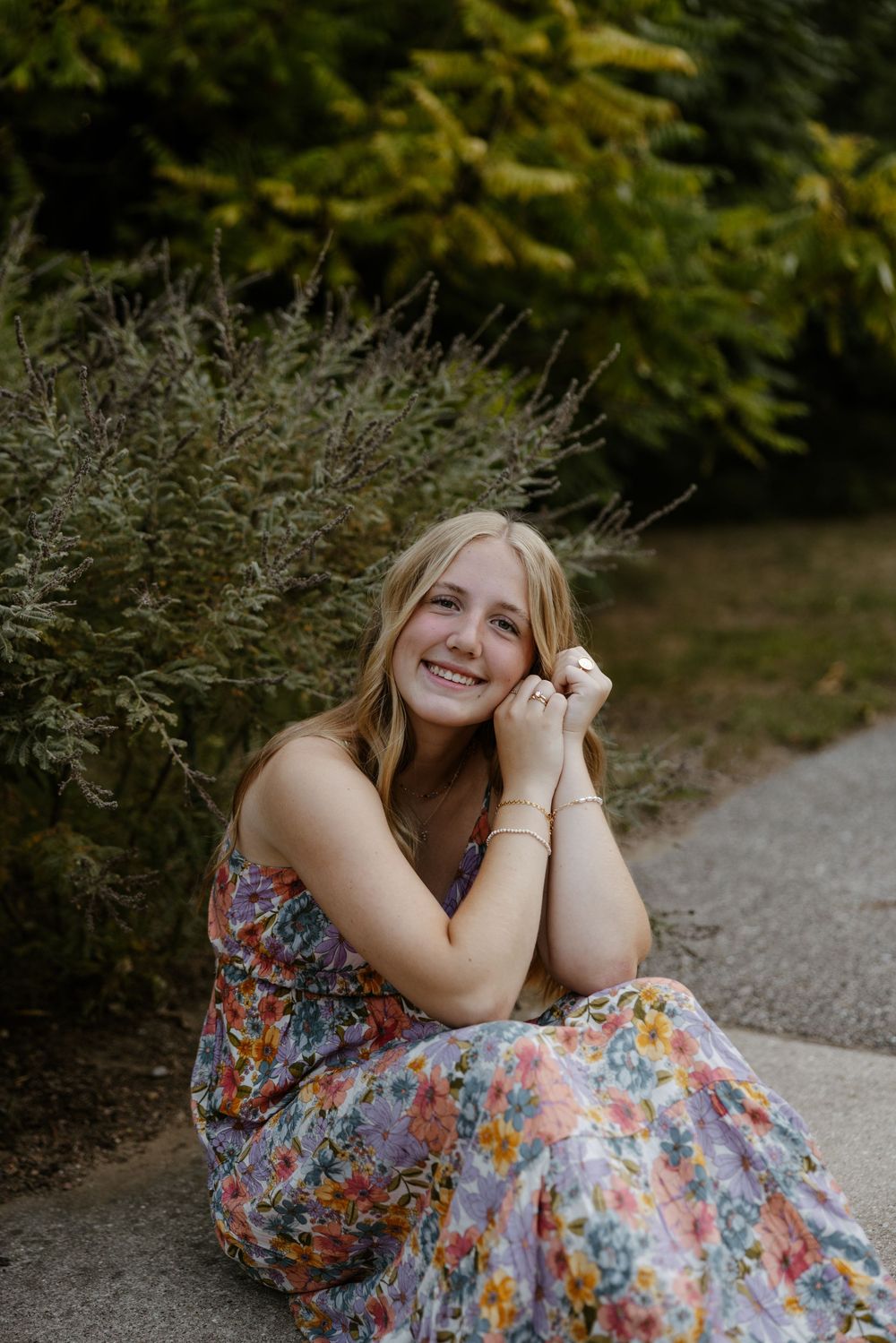 A young woman with long blonde hair sits on a gravel path, surrounded by lush greenery and trees. Fort Worth Texas senior photographer.