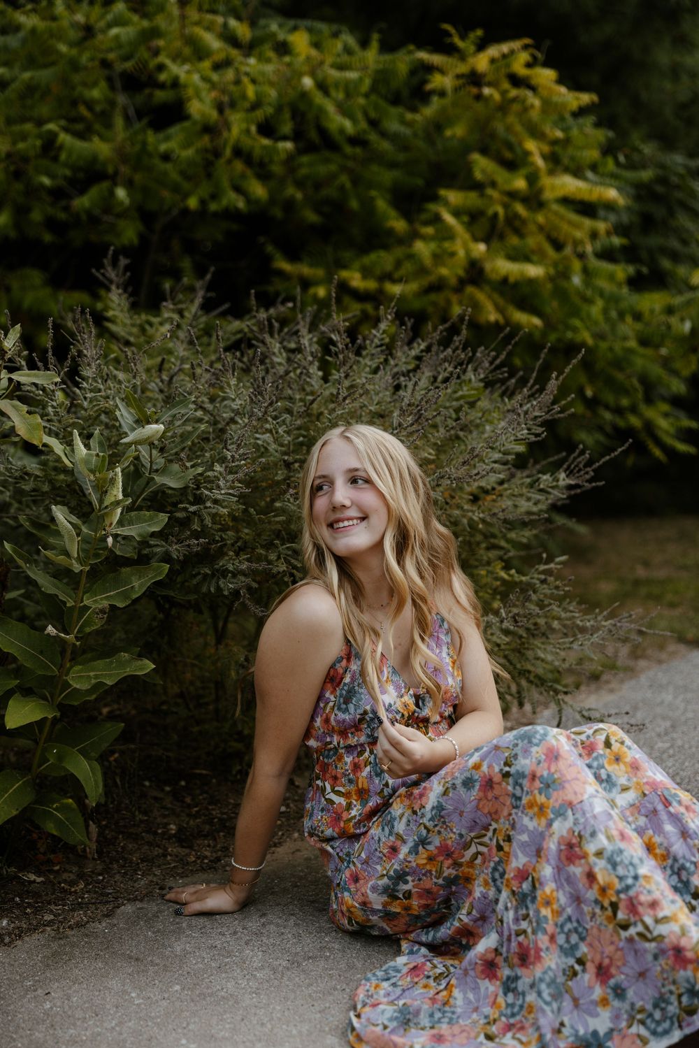 A smiling woman in a colorful floral dress sitting on a path surrounded by lush greenery. Fort Worth Texas senior photographer.