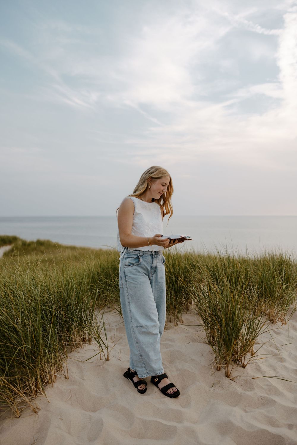 A young woman stands in a grassy dune, holding a mobile device and wearing casual clothing. Fort Worth Texas senior photographer.