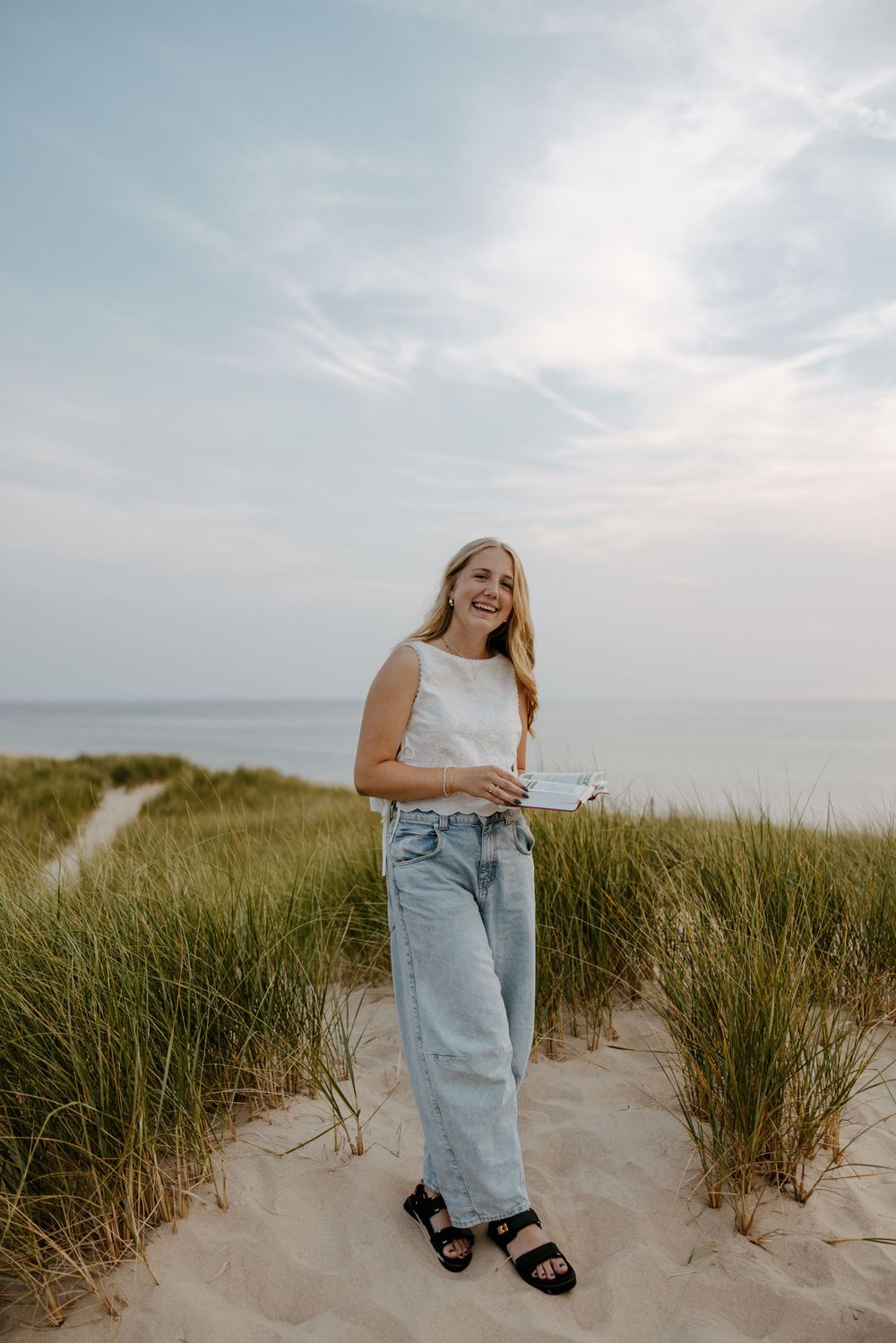 A young woman smiling and standing on a sandy beach with tall grass in the foreground and a body of water in the background. Fort Worth TX